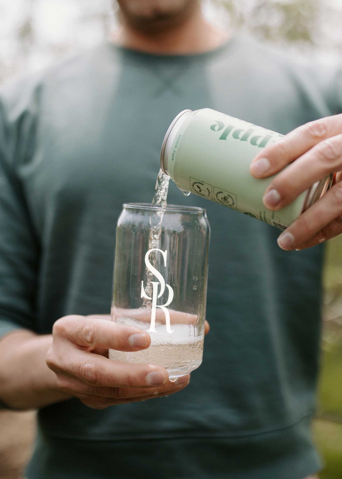 A person outdoors pours a clear liquid from a pale mint green can into a clear glass with a white monogram logo. The person wears a dark green long-sleeve shirt and a black ring, with a softly blurred natural background.