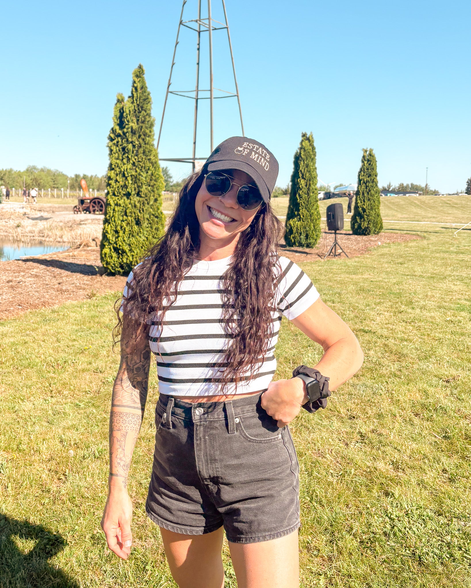 Woman standing outdoors in a grassy area wearing a striped shirt, Estate of Mind hat, sunglasses, and shorts.