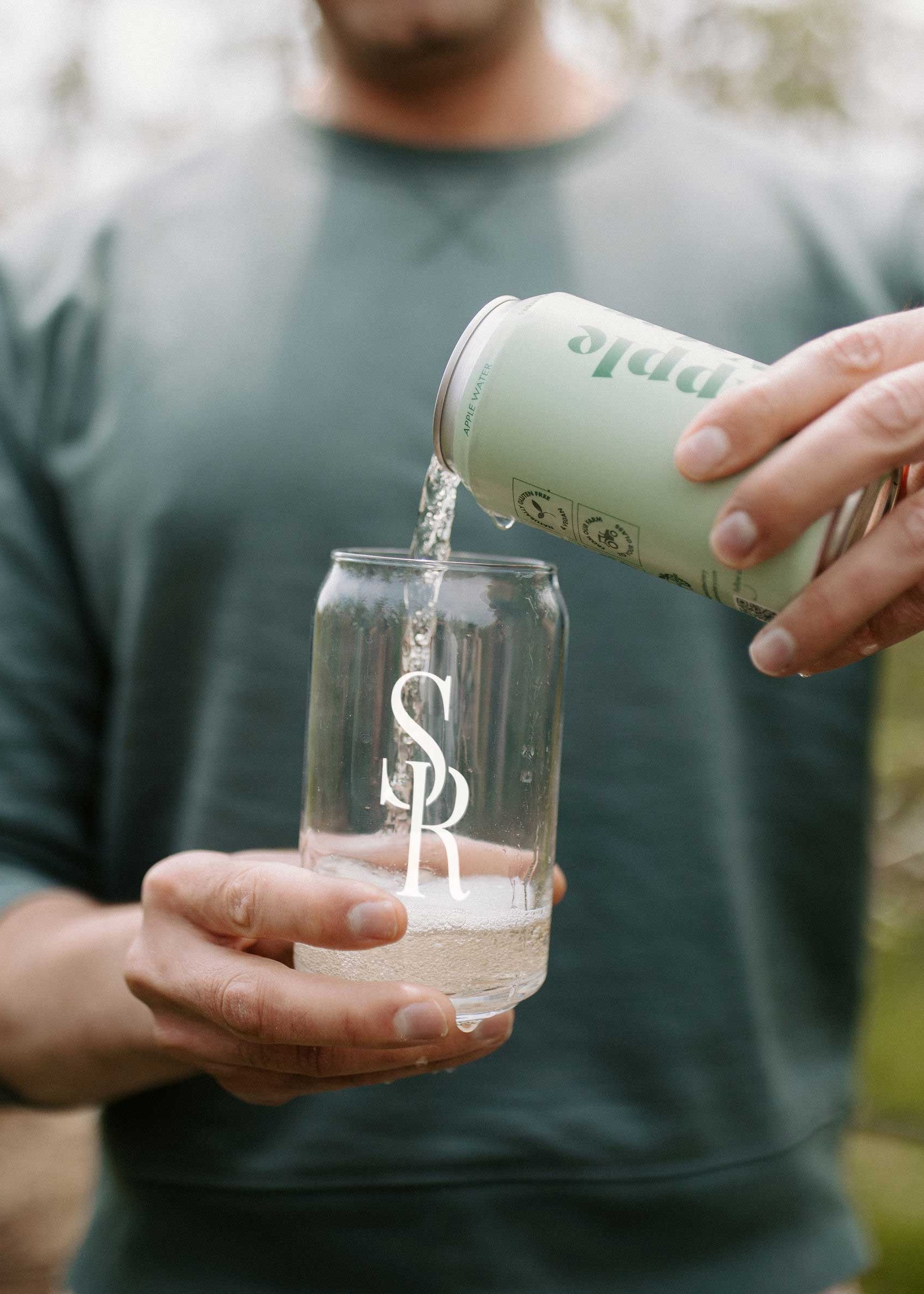 A person outdoors pours a clear liquid from a pale mint green can into a clear glass with a white monogram logo. The person wears a dark green long-sleeve shirt and a black ring, with a softly blurred natural background.