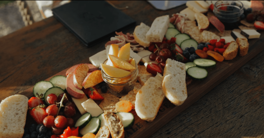 Overhead view of a luxurious, abundant charcuterie board featuring a wide selection of cheese, cured meats, fresh strawberries, grapes, peaches, bread slices, and crackers on a long wooden board.