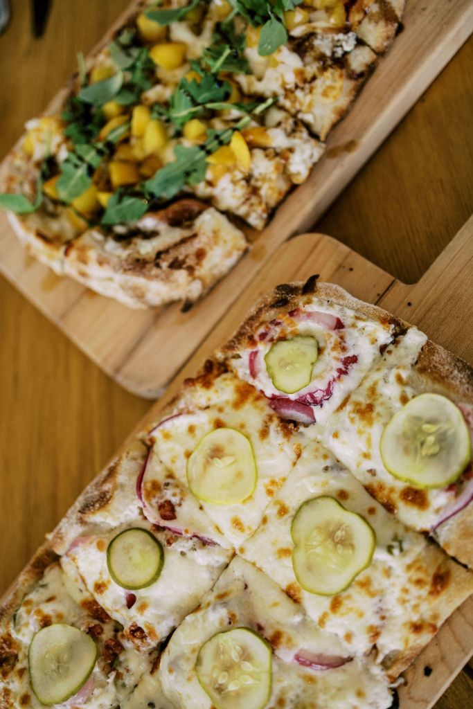 Close-up of two authentic Italian pinsa flatbreads on wooden serving boards: one topped with pickles and red onion, and the other with mango or peach and arugula.