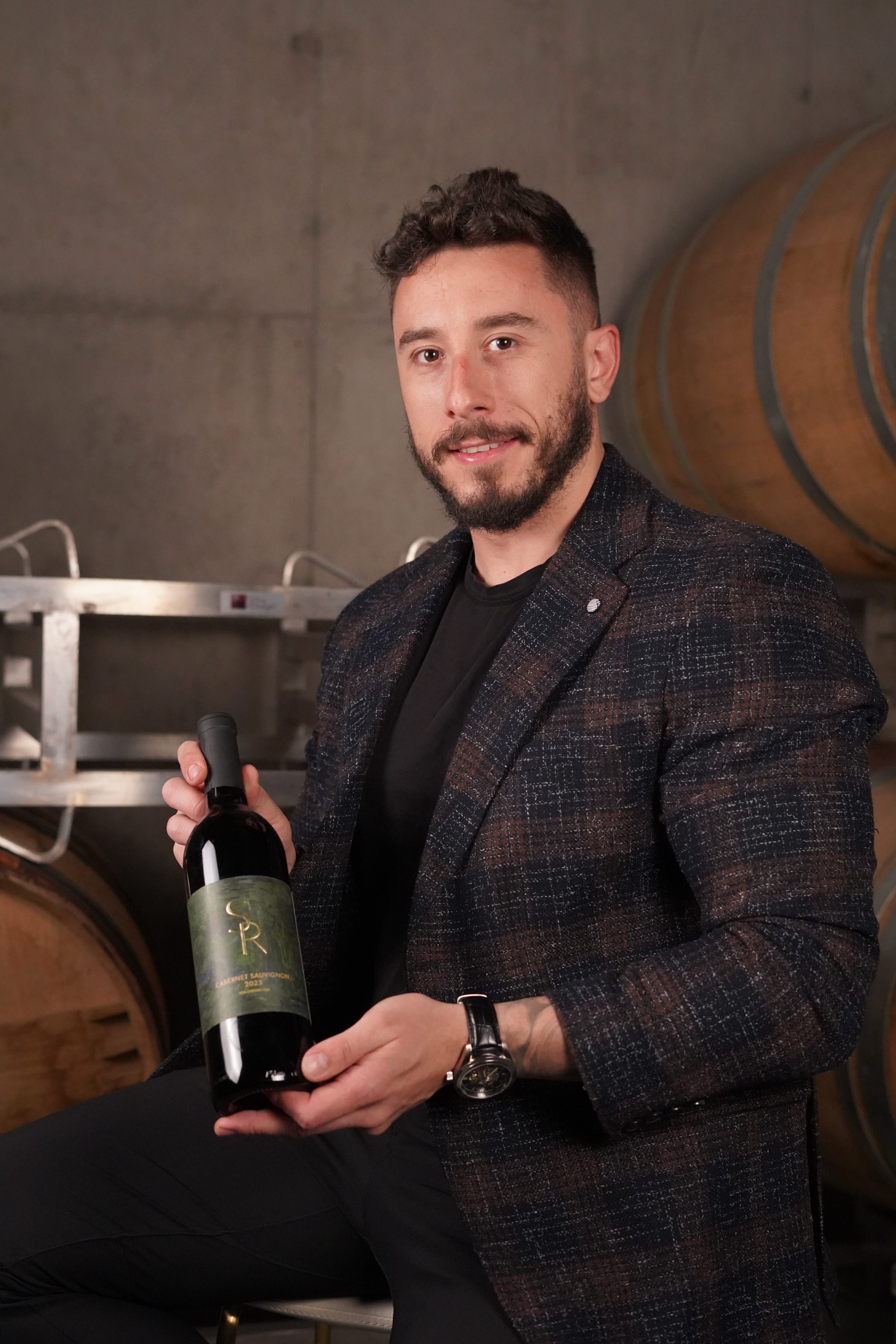 A man holding a bottle of wine while seated in a winery, with barrels in the background.