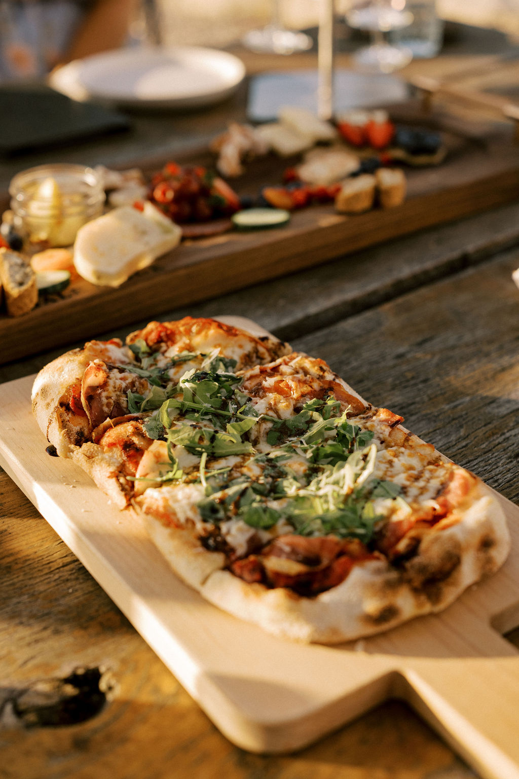 Close-up of a rustic wooden table featuring a freshly baked, long Italian pinsa topped with arugula and cheese, with a charcuterie board visible in the background.