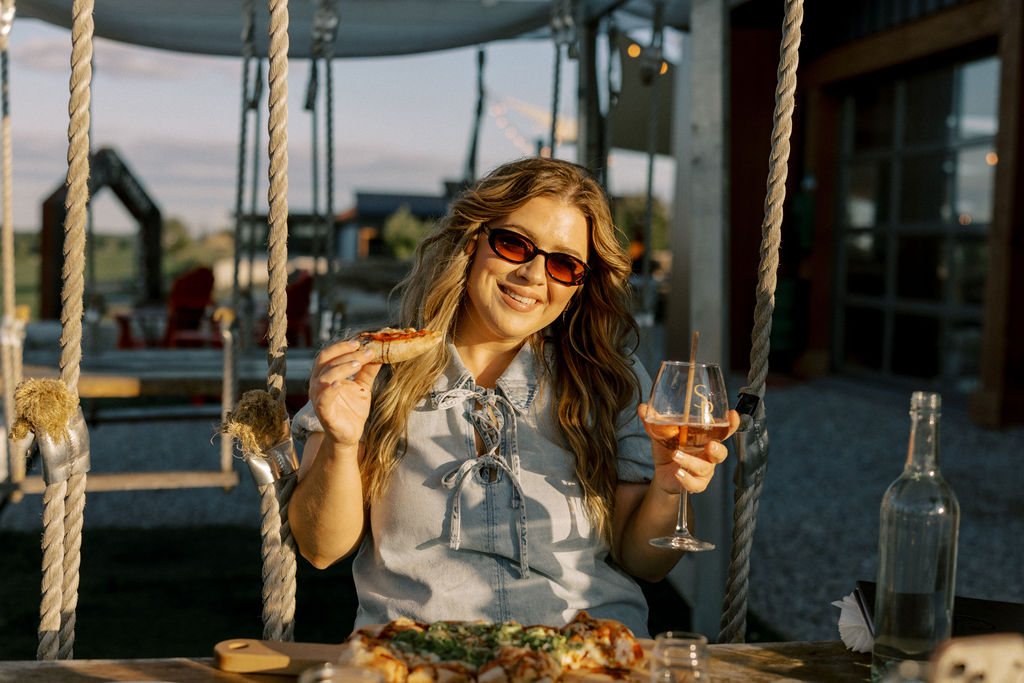 A smiling woman wearing sunglasses, sitting on an outdoor swing table at Shale Ridge, holding a slice of pinsa and a glass of rosé wine in the warm afternoon sun.