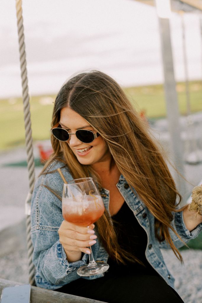 A young woman in sunglasses and a denim jacket smiling while sitting on a swing table at Shale Ridge Estate, holding a glass of a refreshing signature cocktail or wine.