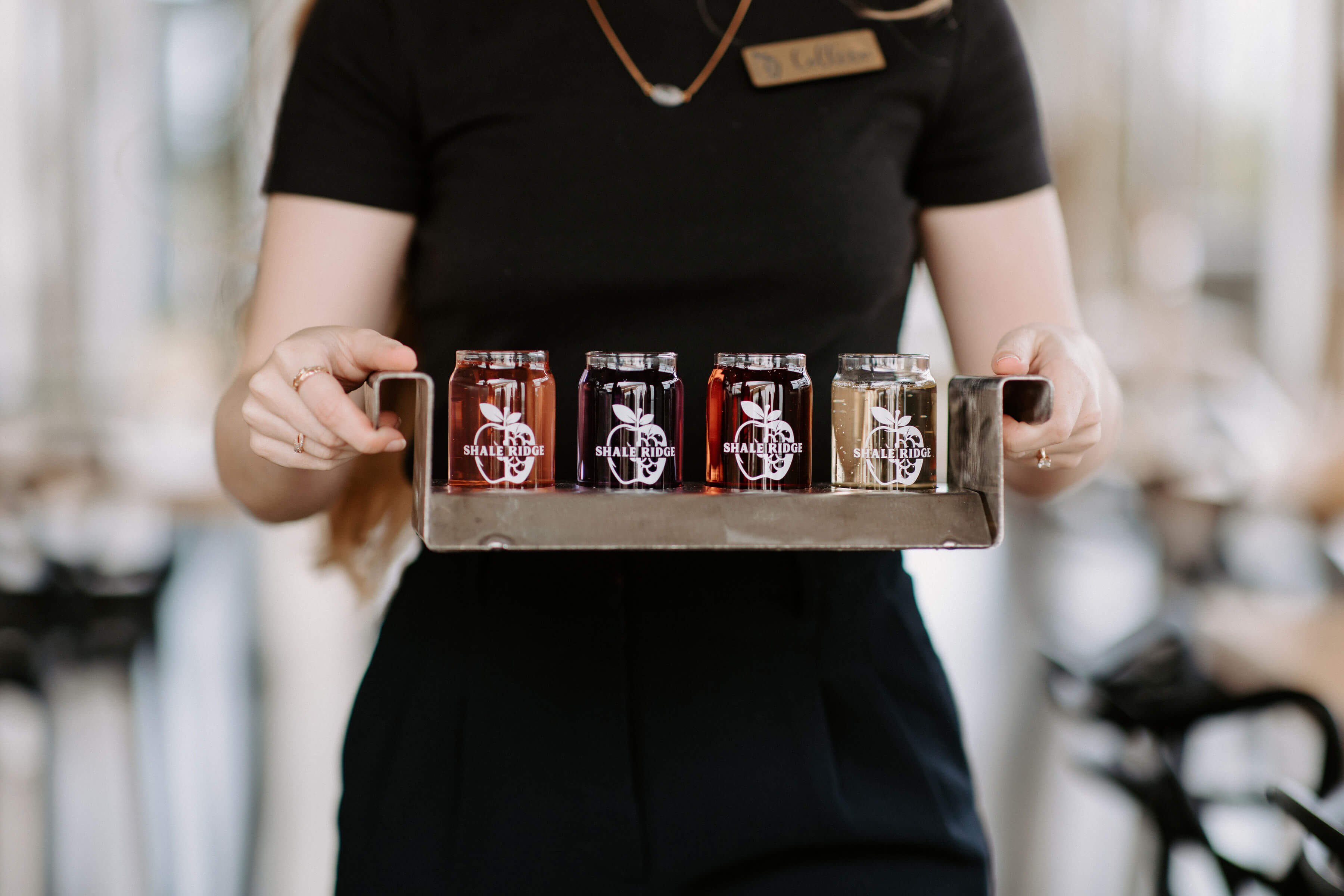 A Shale Ridge staff member in a black uniform holding a metal tray with four sample glasses of brightly colored craft cider, each featuring the Shale Ridge logo.
