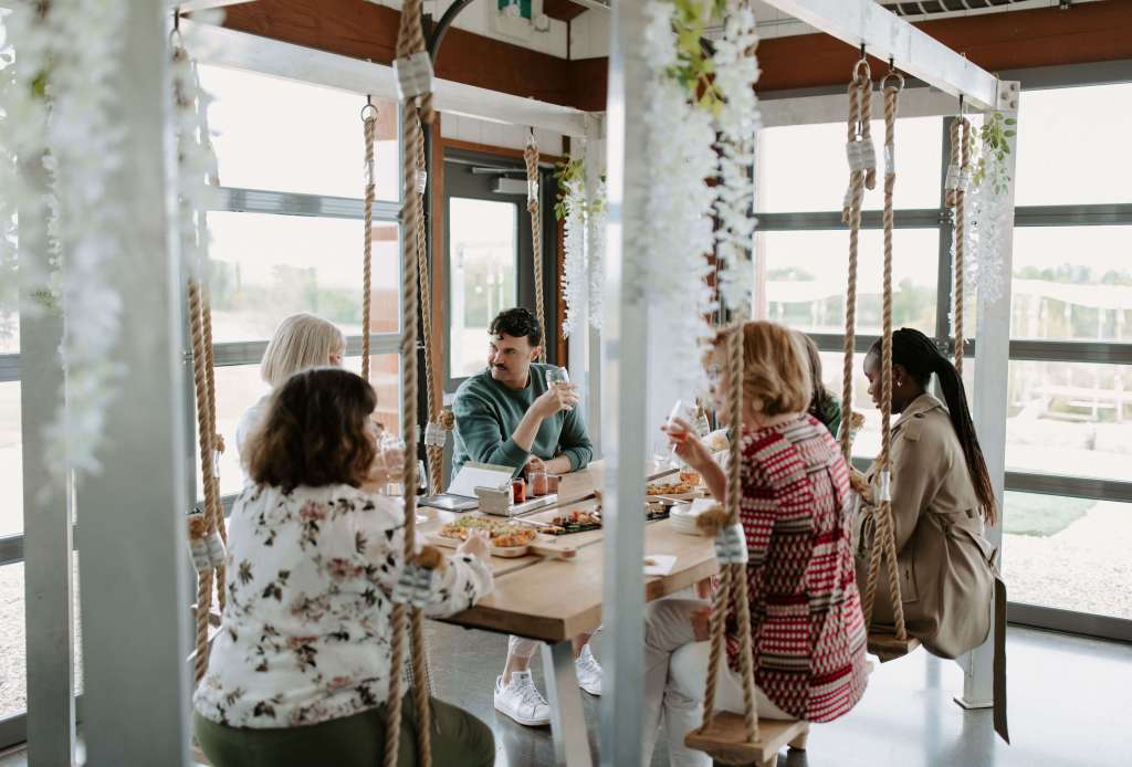 A group of six people sitting on hanging rope swing seats around a wooden table, enjoying lunch and drinks inside a bright, modern event space at Shale Ridge.