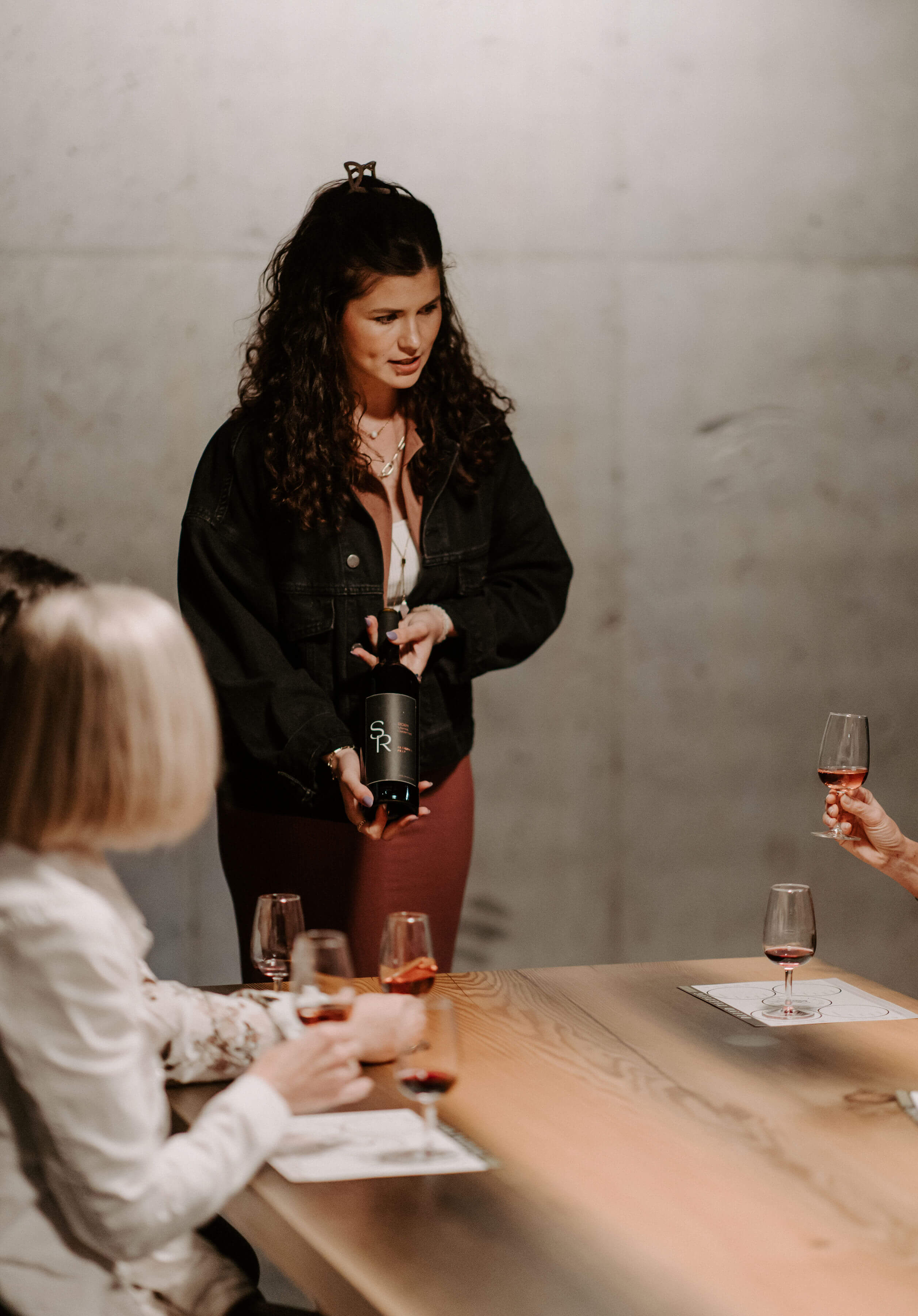A woman with curly hair holding a bottle of wine in a tasting room, surrounded by a group of people engaged in conversation with wine glasses on the table.