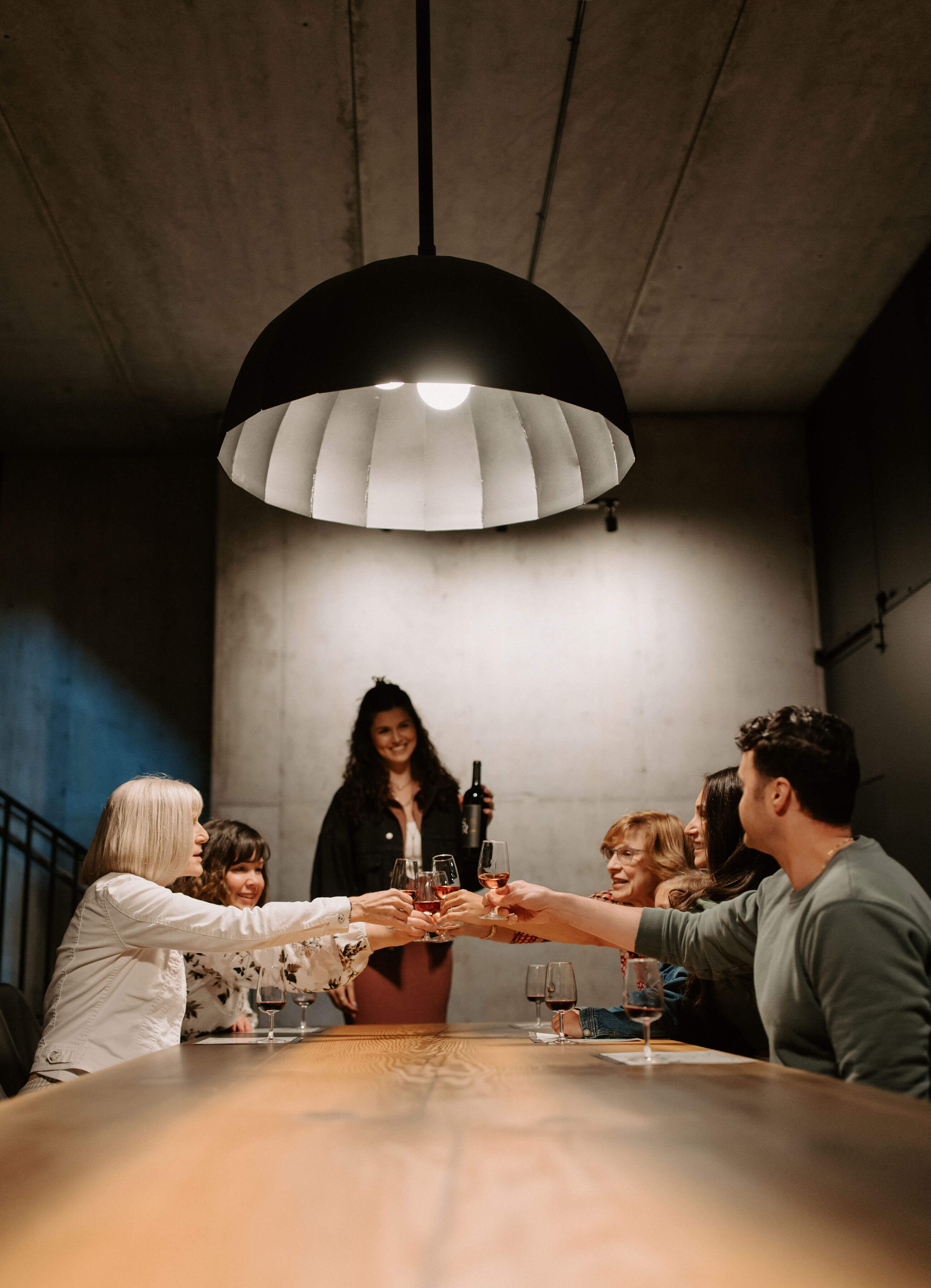 A group of friends seated at a long wooden table beneath a large black pendant light, raising glasses of red wine in a toast, led by a smiling server holding a bottle in a modern, dimly lit tasting room.