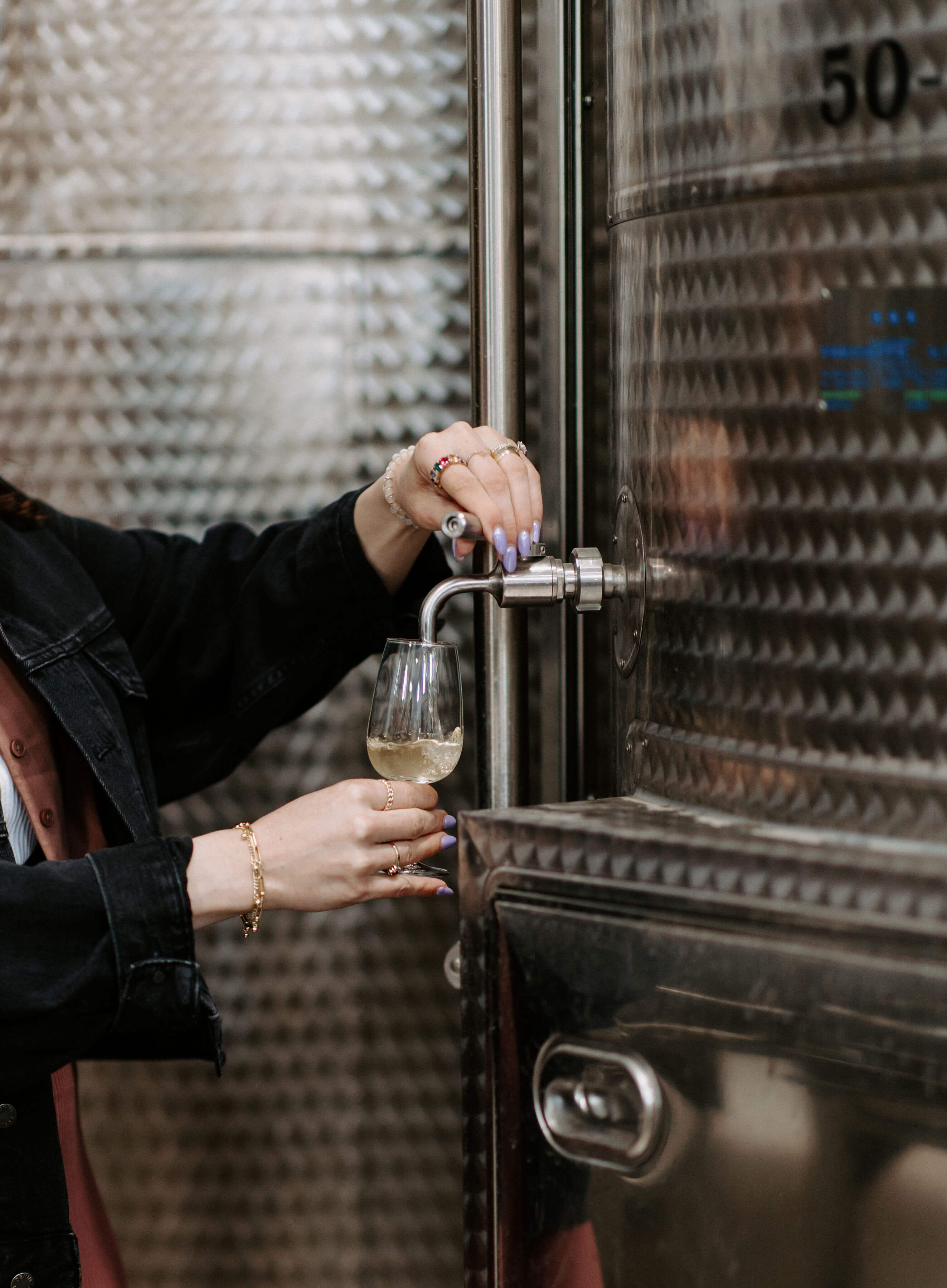 A person pouring white wine into a glass from a stainless steel wine fermentation tank.
