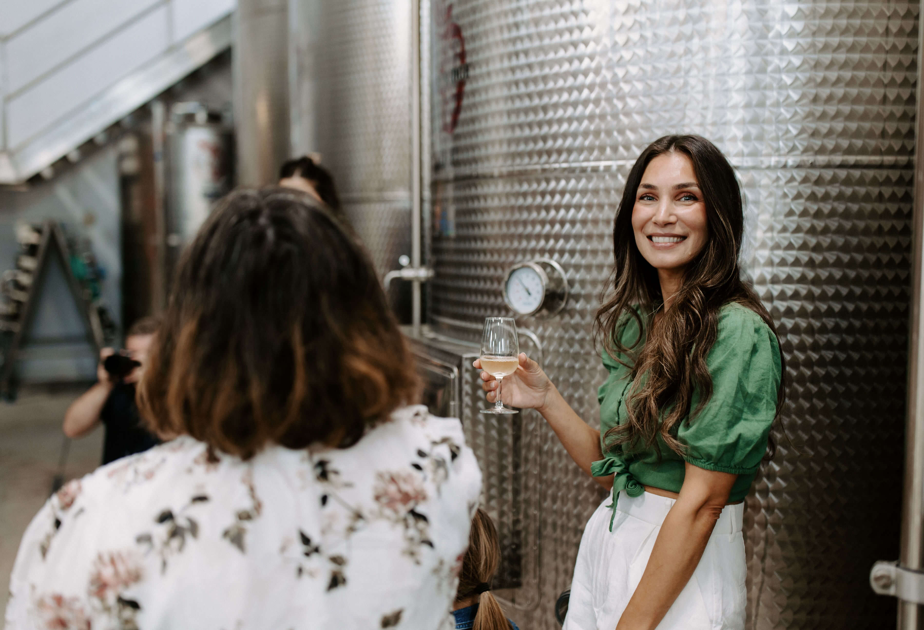Woman holding a glass of wine while smiling, standing in a winery with metallic fermentation tanks in the background.
