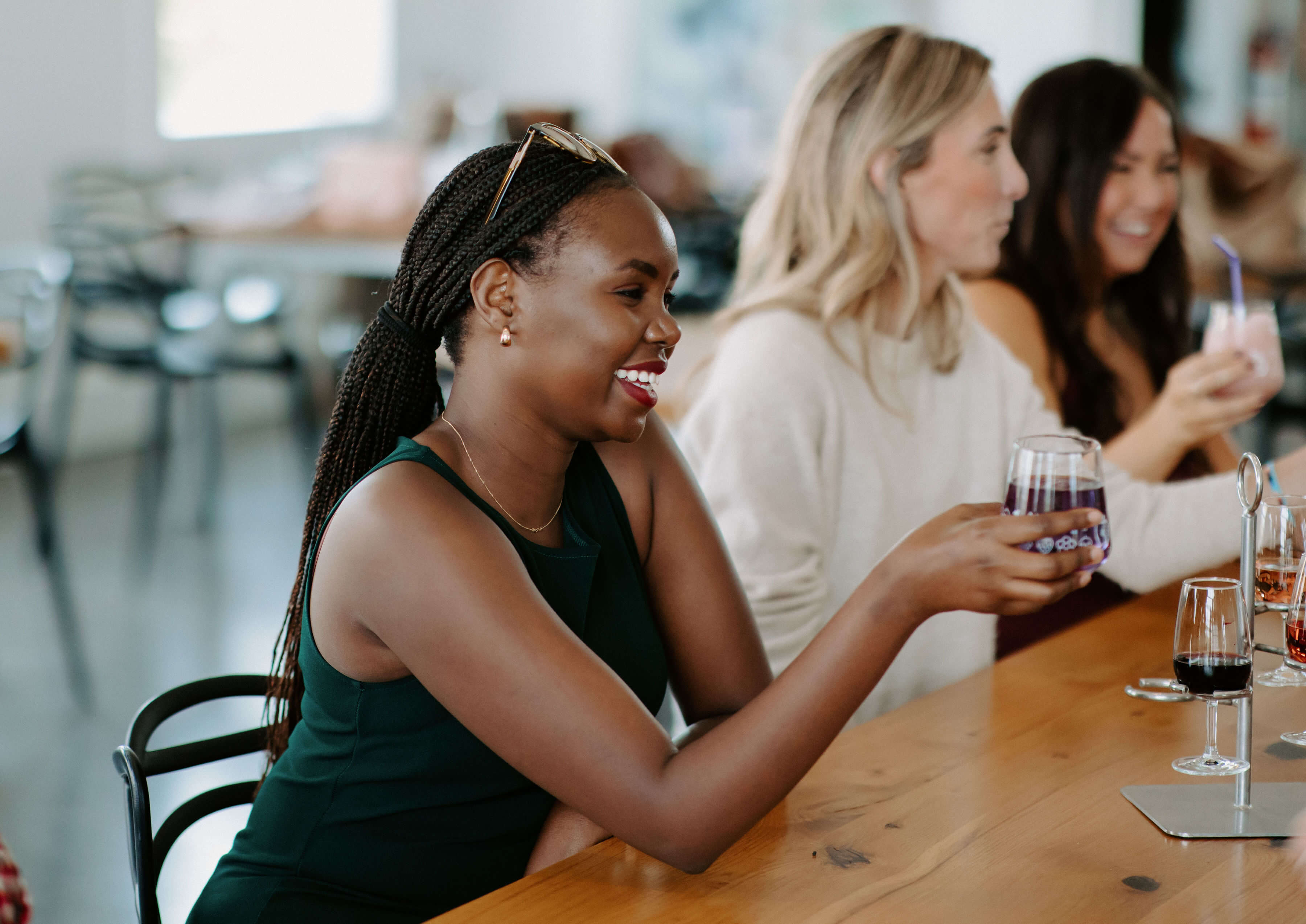 A smiling woman holding a glass of purple wine or cider while seated at a wooden bar, with two friends holding drinks blurred in the background, capturing a cheerful tasting event.