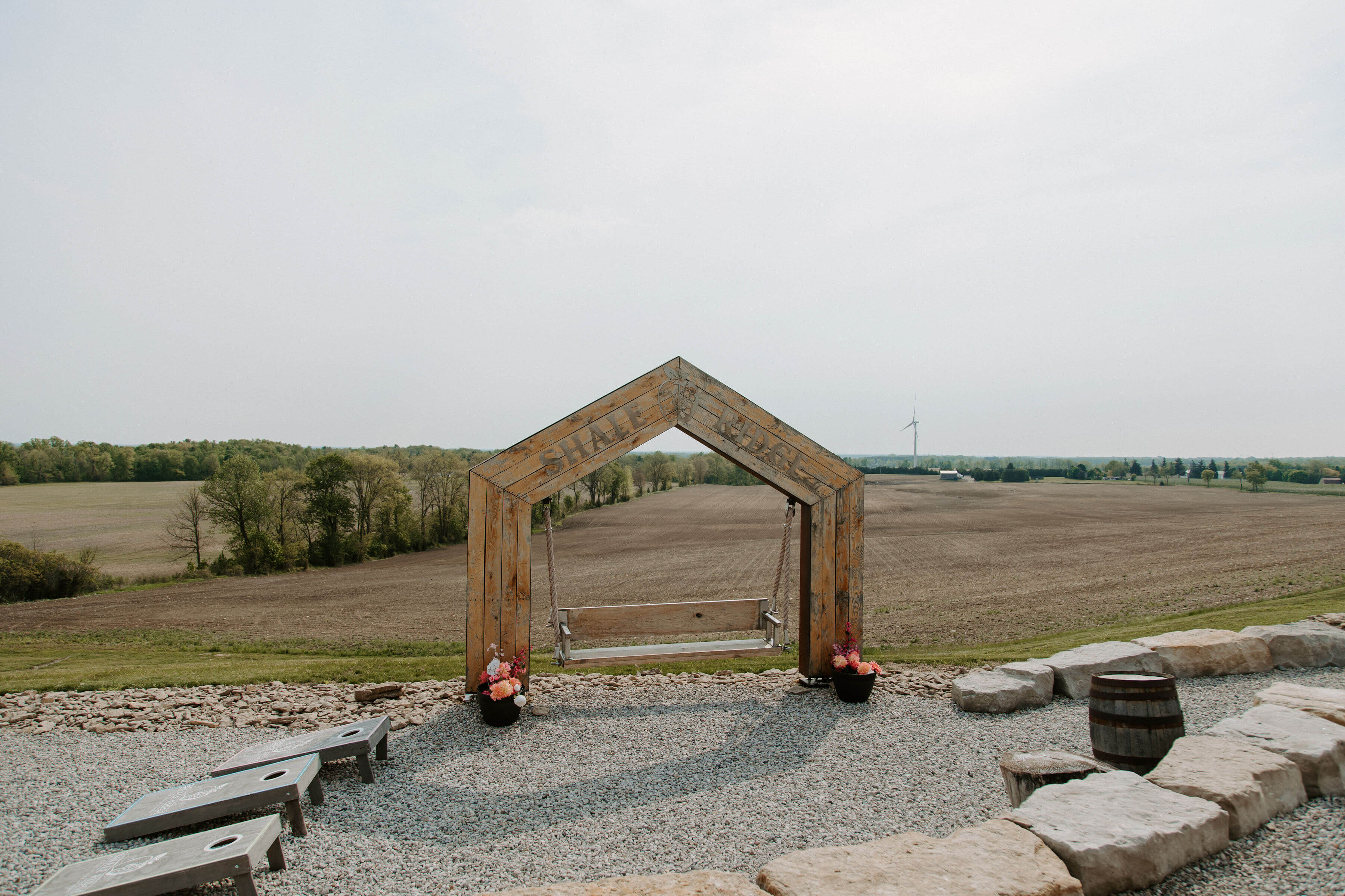 The Shale Ridge outdoor patio featuring a wooden, house-shaped frame with a swing, situated on a stone patio with cornhole games, facing expansive farmland and a distant wind turbine.