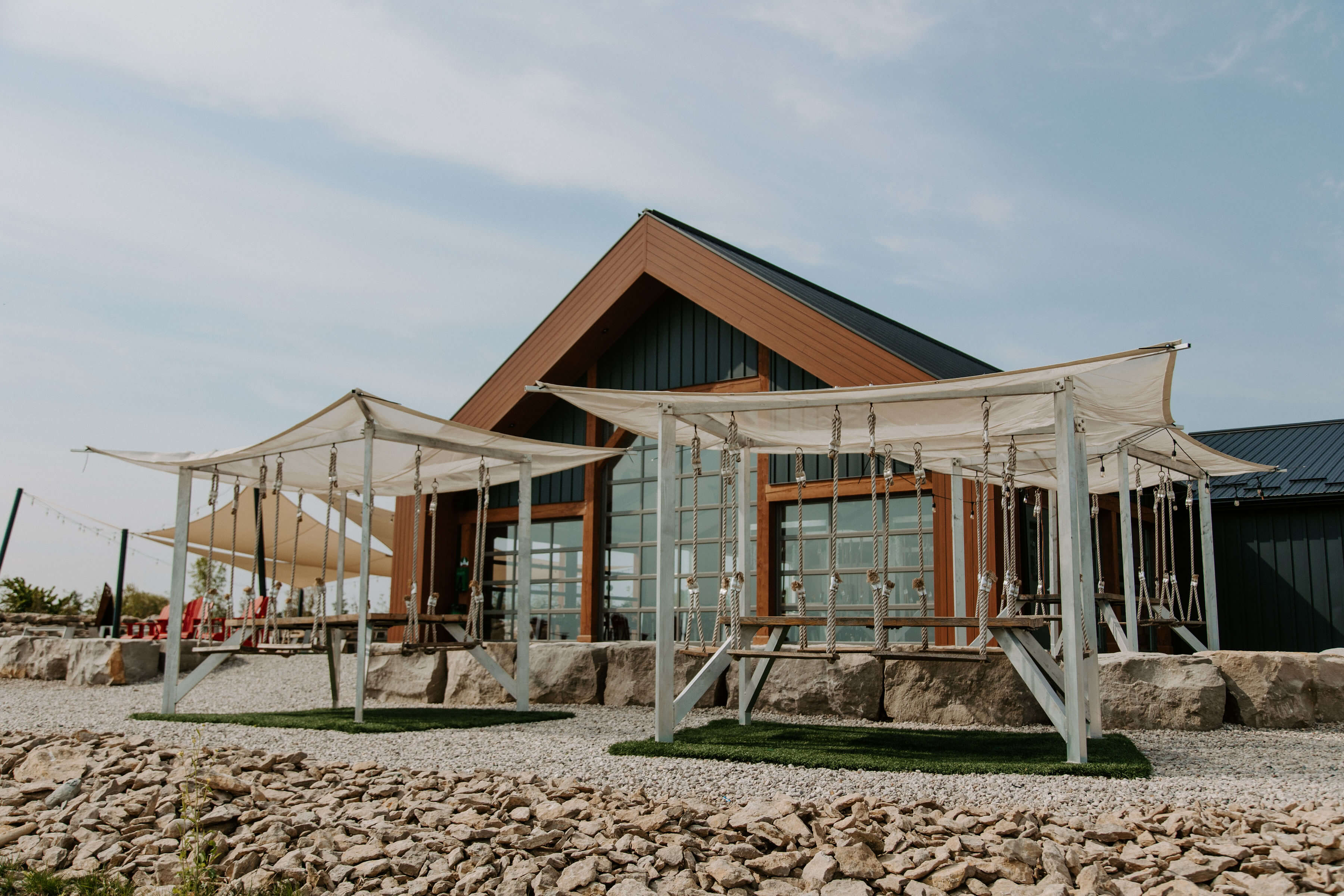 Exterior view of a modern building with large windows and a triangular roof, featuring swings under canopies in a gravel area.