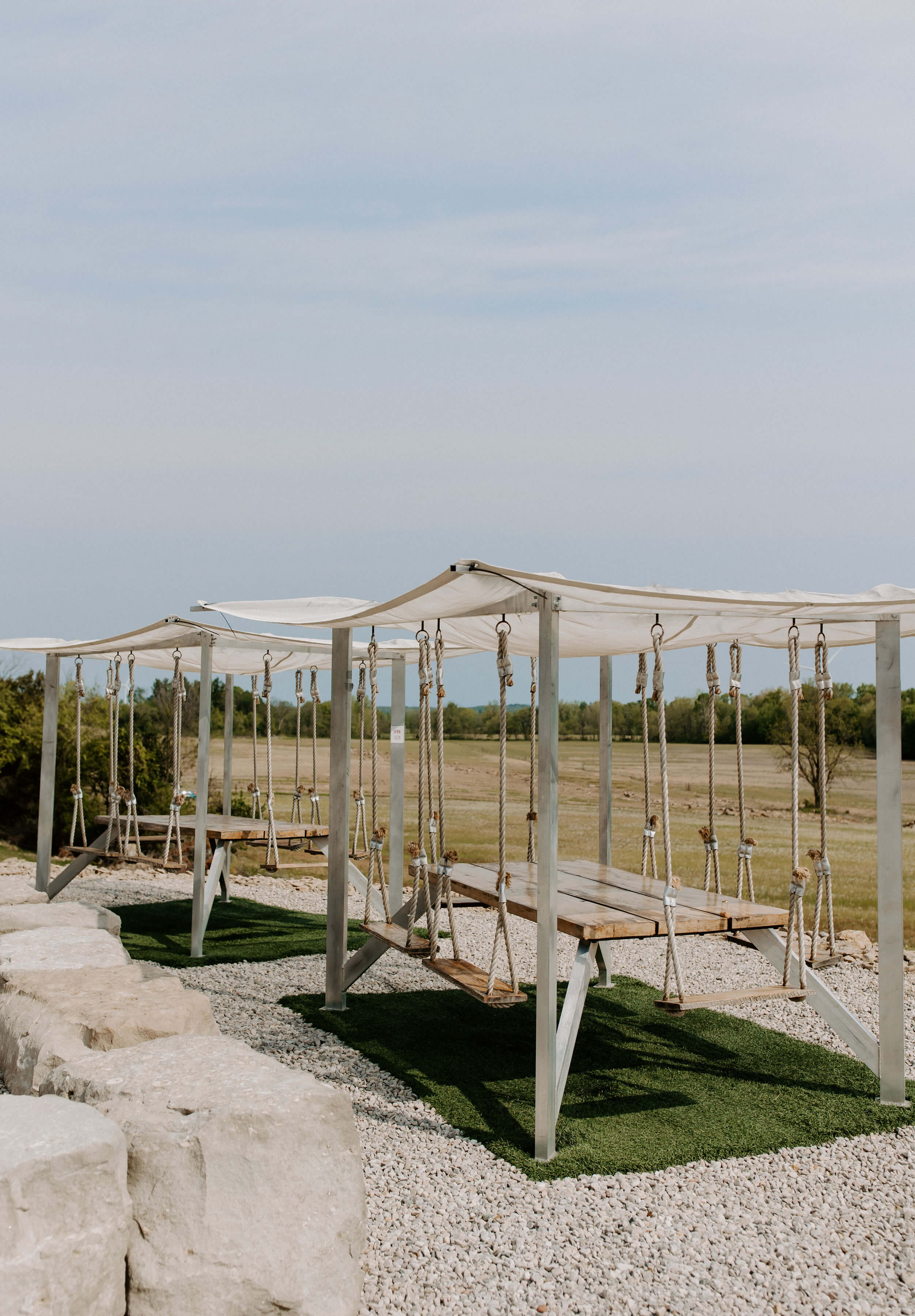 Two unique outdoor swing tables with wooden tabletops, rope seating, and white canopies, set over artificial grass and crushed stone on the Shale Ridge patio, overlooking the countryside.