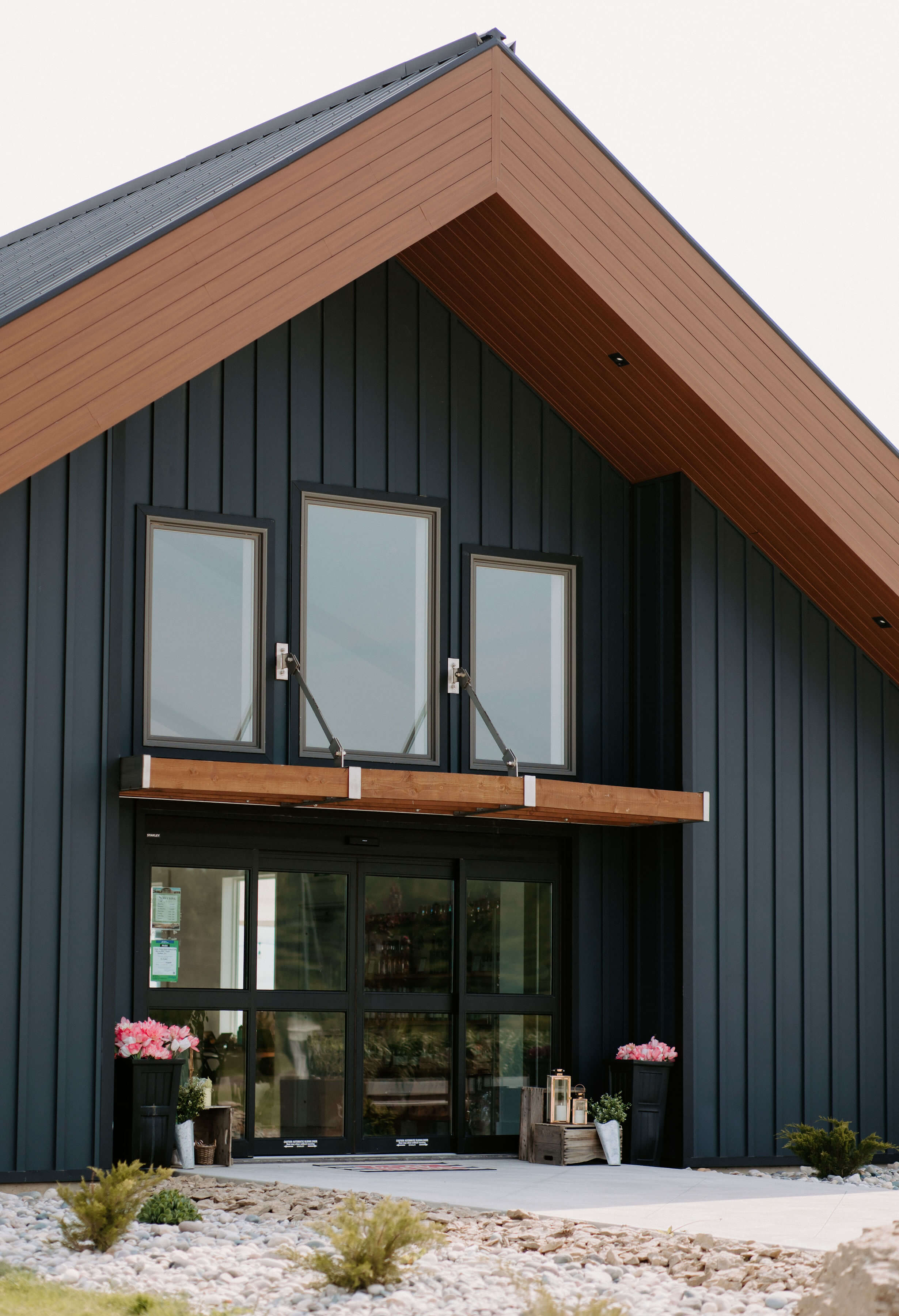 Exterior view of a modern building with a sloped roof, featuring large windows and a welcoming entrance adorned with potted flowers.