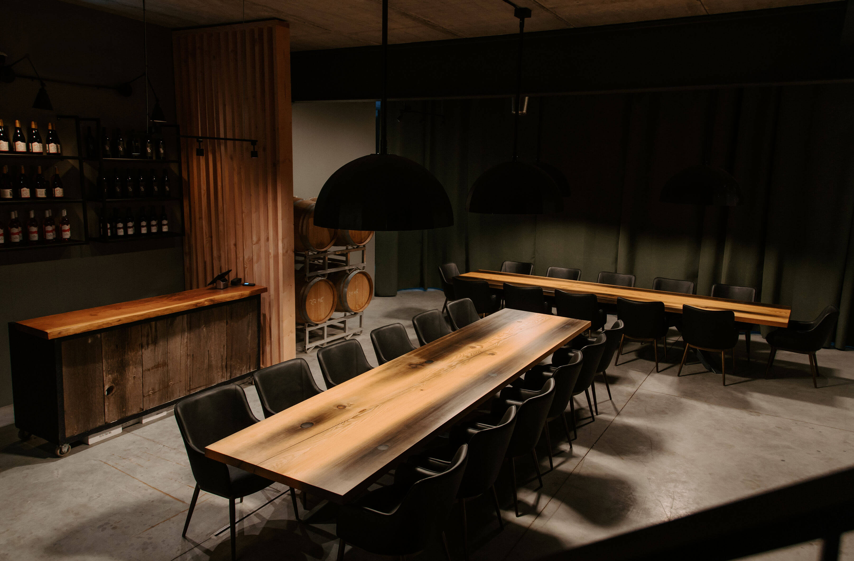 title The modern, dimly lit private event room at Shale Ridge Winery featuring two long, rustic wooden tables with black chairs, wine barrels, a wooden bar console, and minimalist pendant lighting.