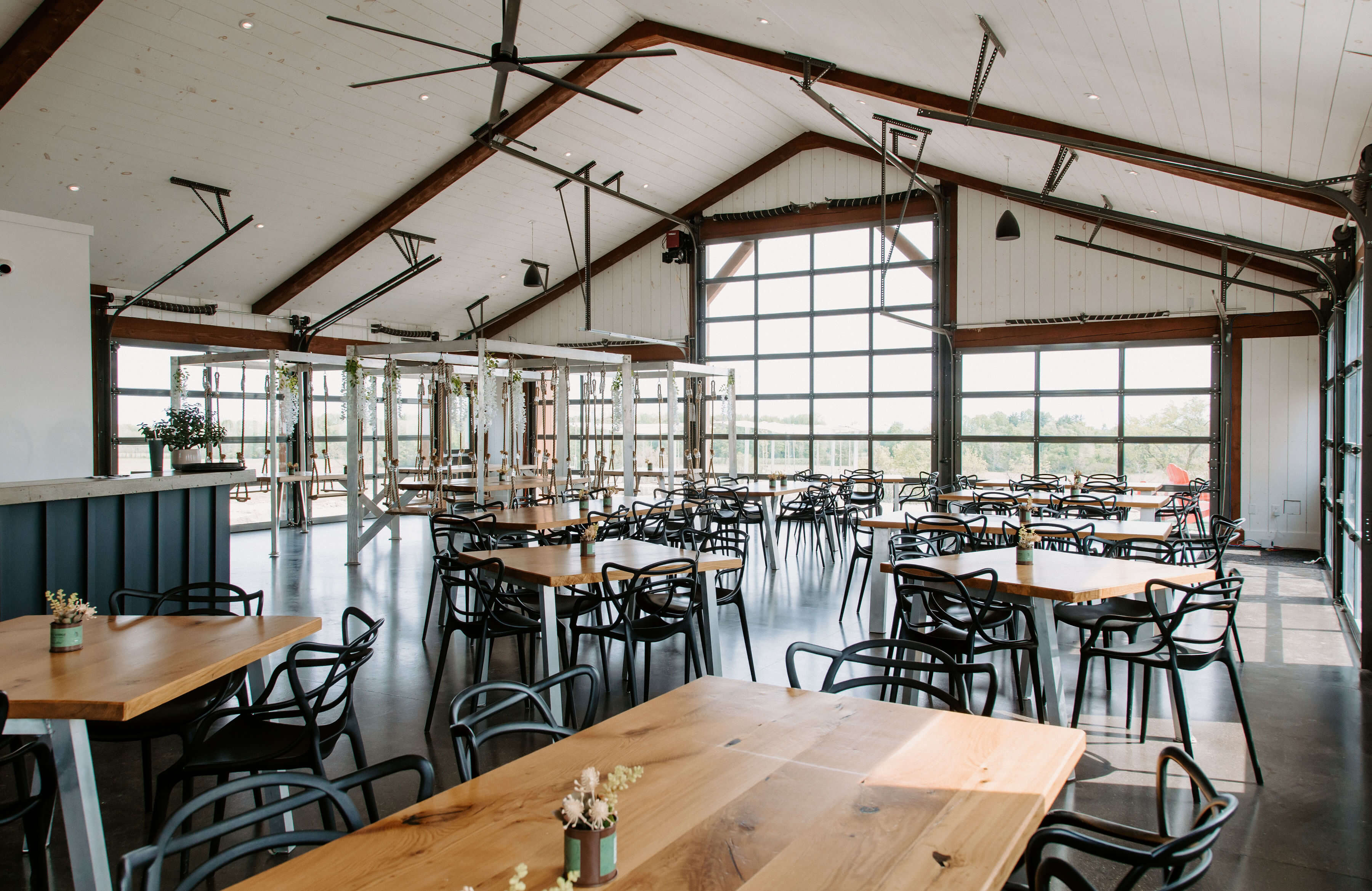 The spacious, light-filled Shale Ridge tasting room featuring a high vaulted white ceiling, exposed wooden beams, multiple light wood tables with black chairs, and large industrial glass garage doors overlooking the vineyard.