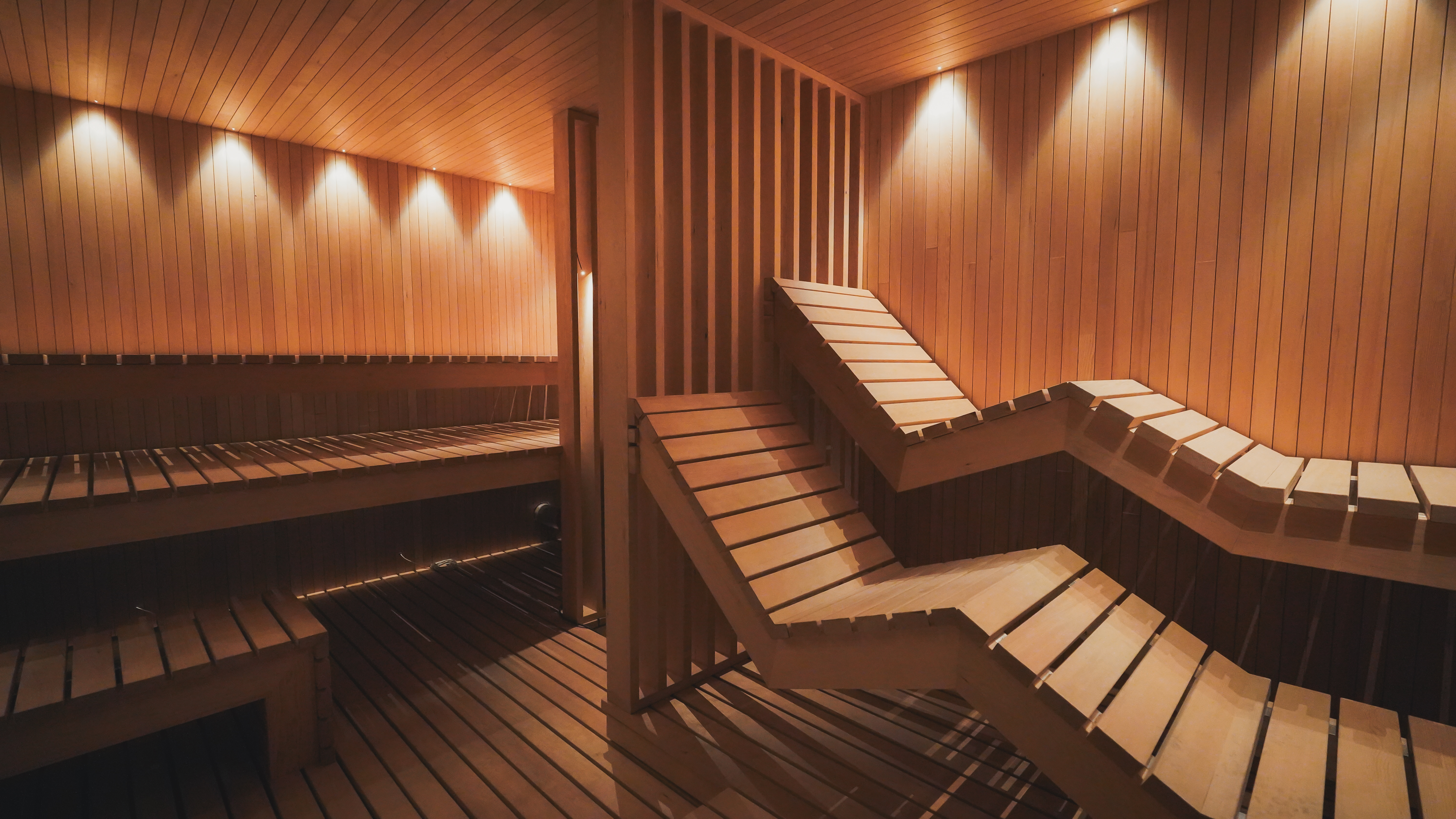 Interior of a luxury modern sauna with tiered wooden seating, recessed lighting, and a geometric wood divider.