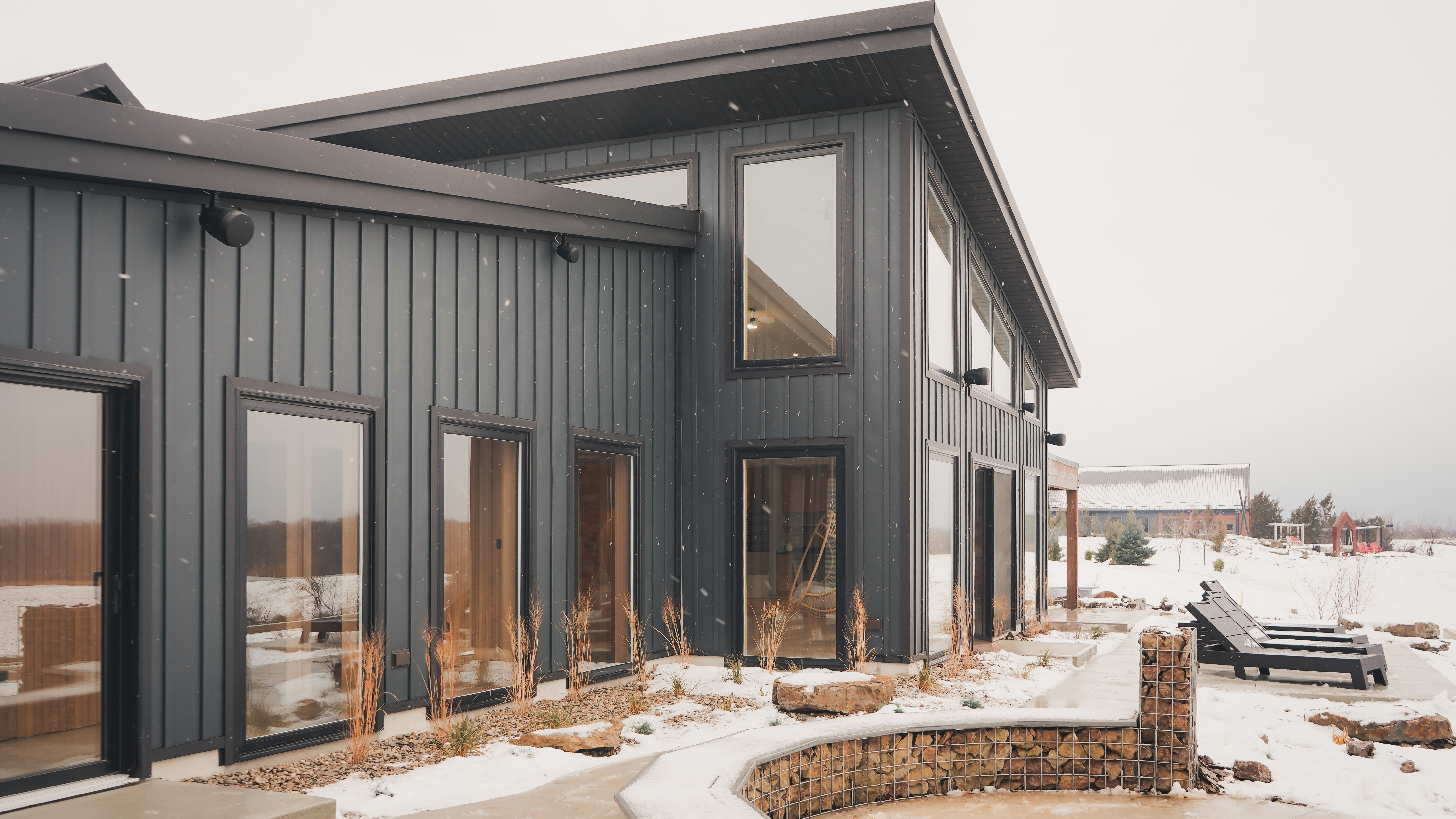 Modern dark charcoal grey building with large windows and a gabion stone wall during a light snowfall.
