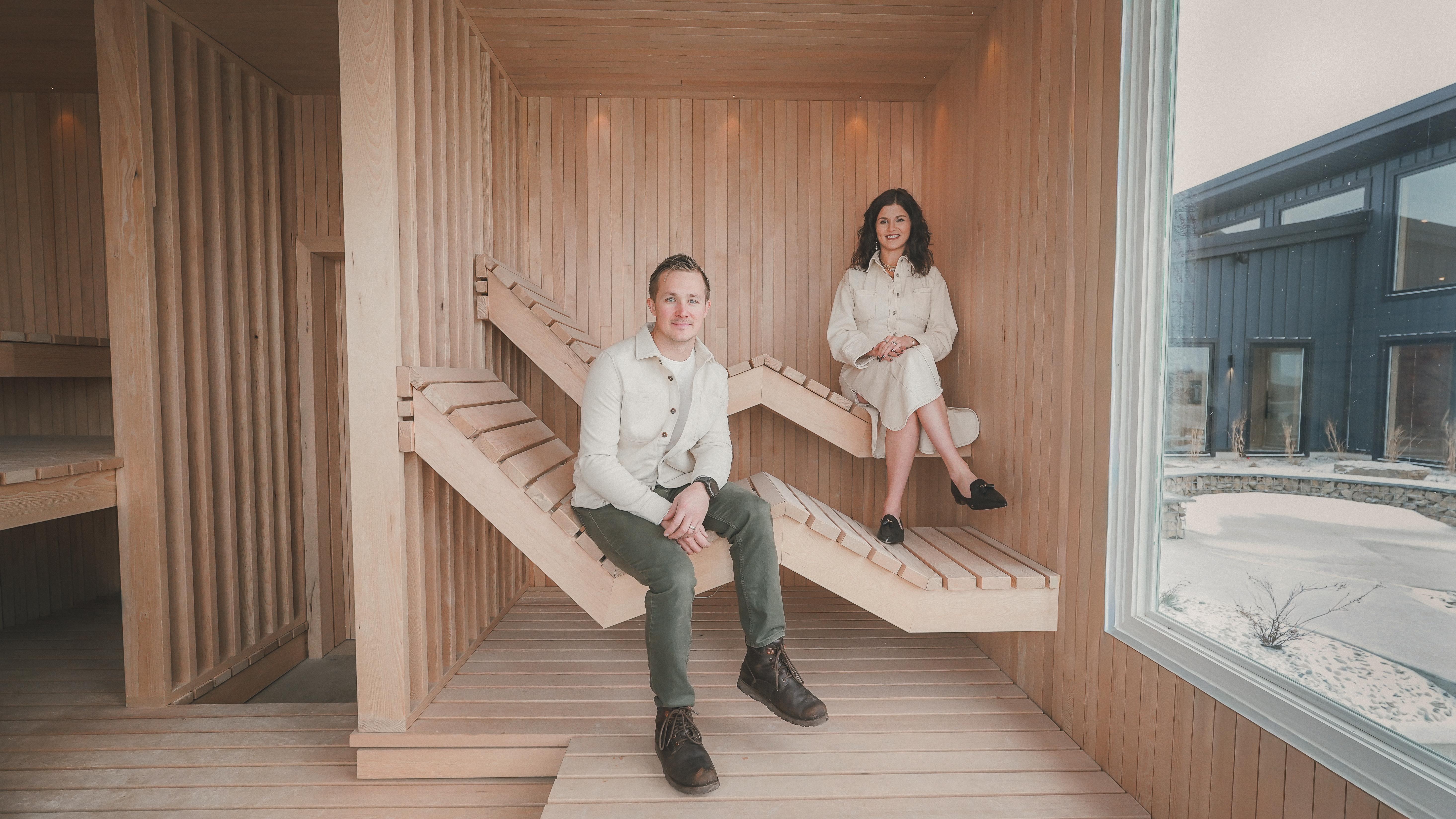 Man and woman sitting on modern, geometric wooden sauna loungers in a sunlit, wood-paneled room.