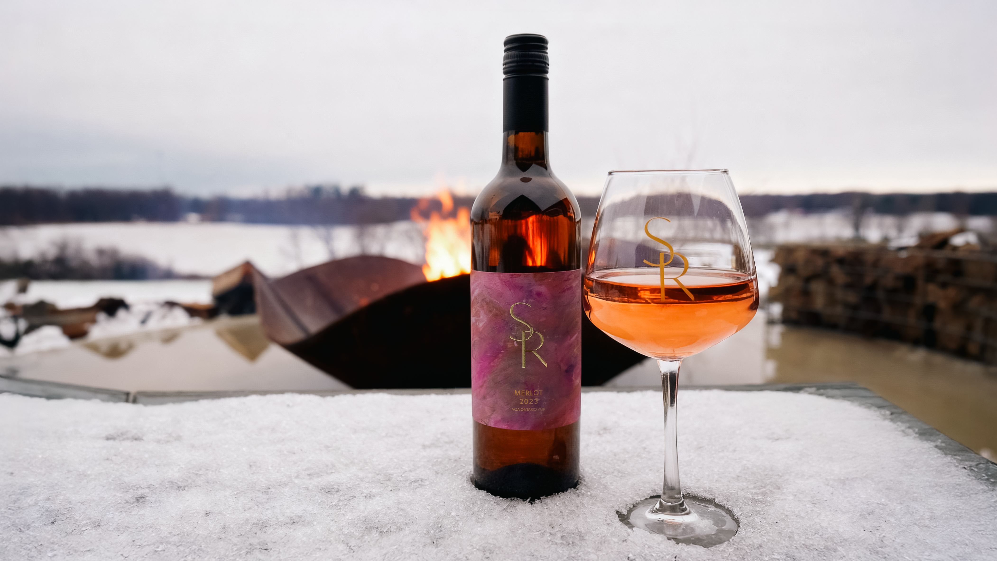 Bottle of Shale Ridge Merlot Rosé and a glass of rosé wine on a snow-covered surface with an outdoor fire pit burning in the background.