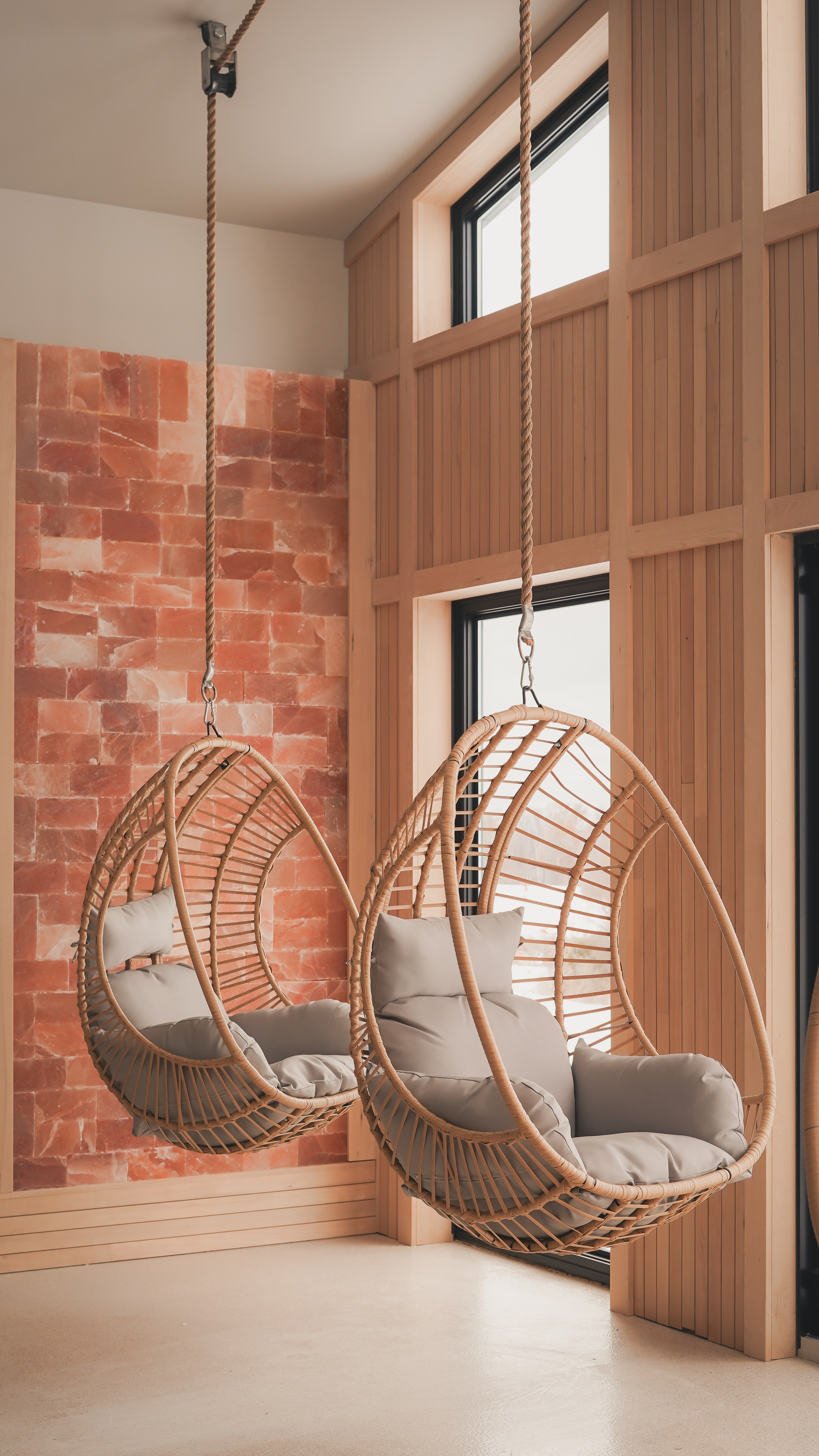 Two wicker egg chairs with grey cushions hanging in front of a Himalayan salt brick wall and floor-to-ceiling windows.