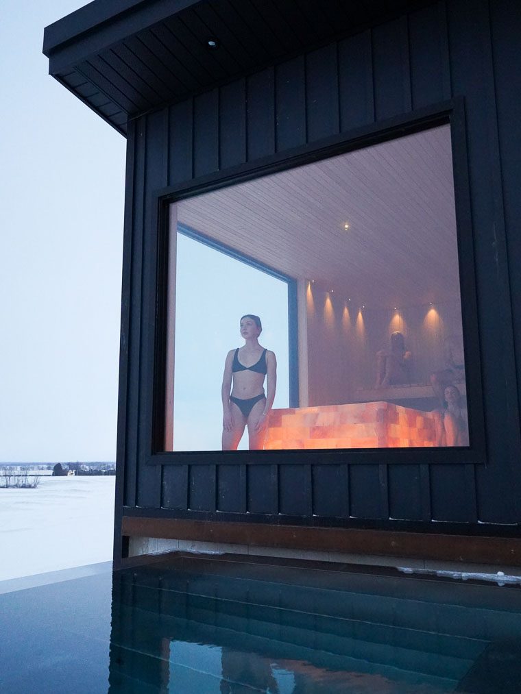 title A woman in a bikini stands in front of a large window, looking out from a modern building with a sauna. The exterior is black, and the surrounding landscape is covered in snow. A serene pool reflects the building and the snowy surroundings.
