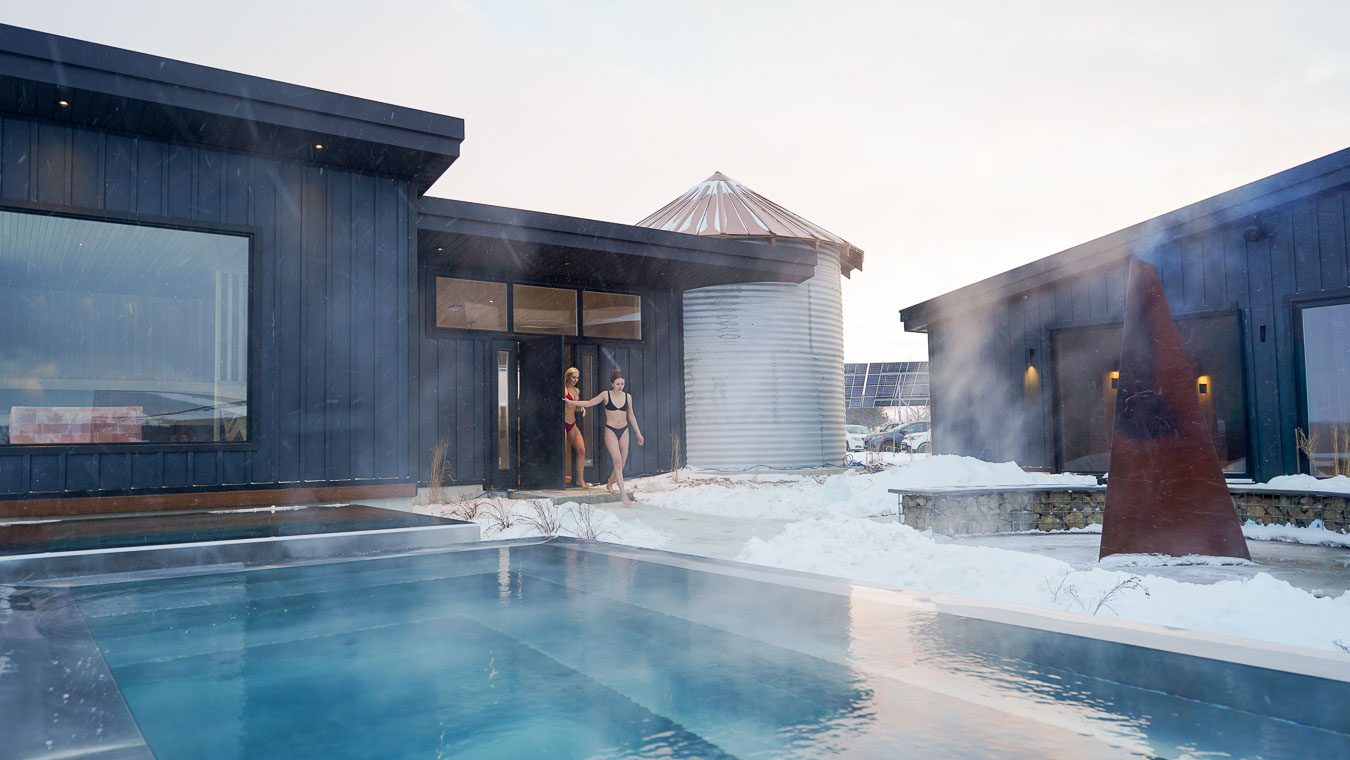 Two women exit a modern building wearing swimwear, with a snow-covered landscape and a steaming pool in the foreground.